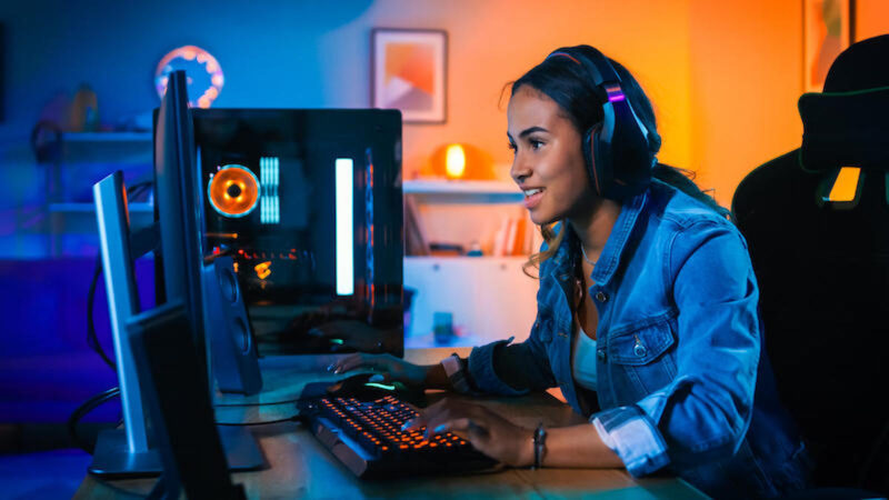 Pretty and excited black gamer girl in headphones is playing first person shooter online video game on her computer. Room and pc have colorful neon led lights. Cozy evening at home.